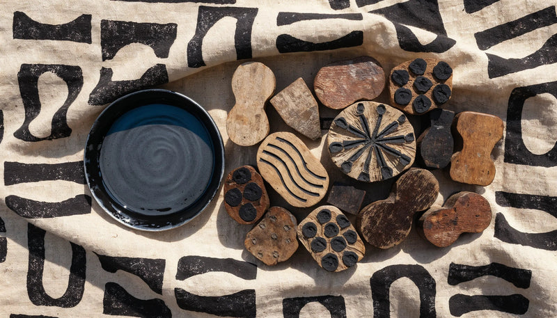 Hand carved wooden printing stamps on undyed cotton cloth beside a dish of dark ink, with geometric motifs stamped in rows