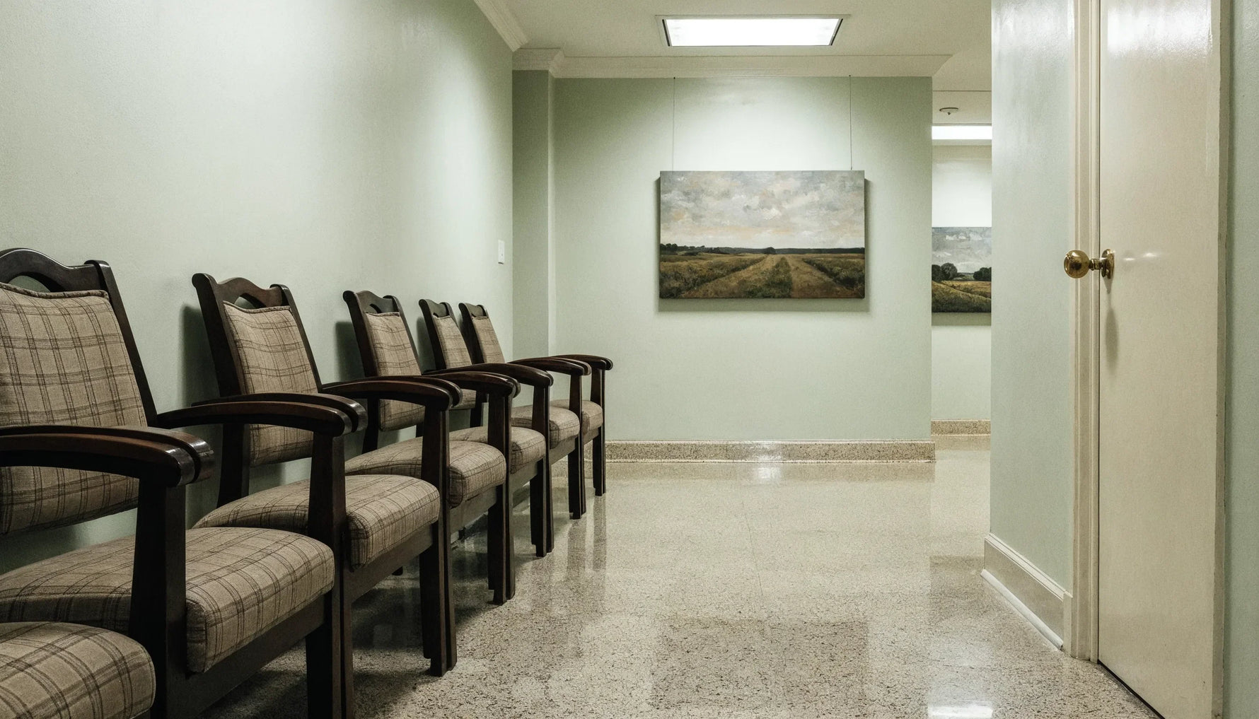 A quiet waiting room with upholstered chairs facing a mid contrast landscape canvas hung across the room, lit softly from above.