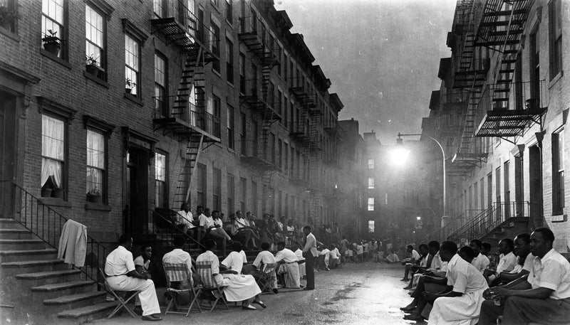 Black and white photograph of a 1930s city block at night with neighbors gathered on brownstone stoops under a single streetlamp.