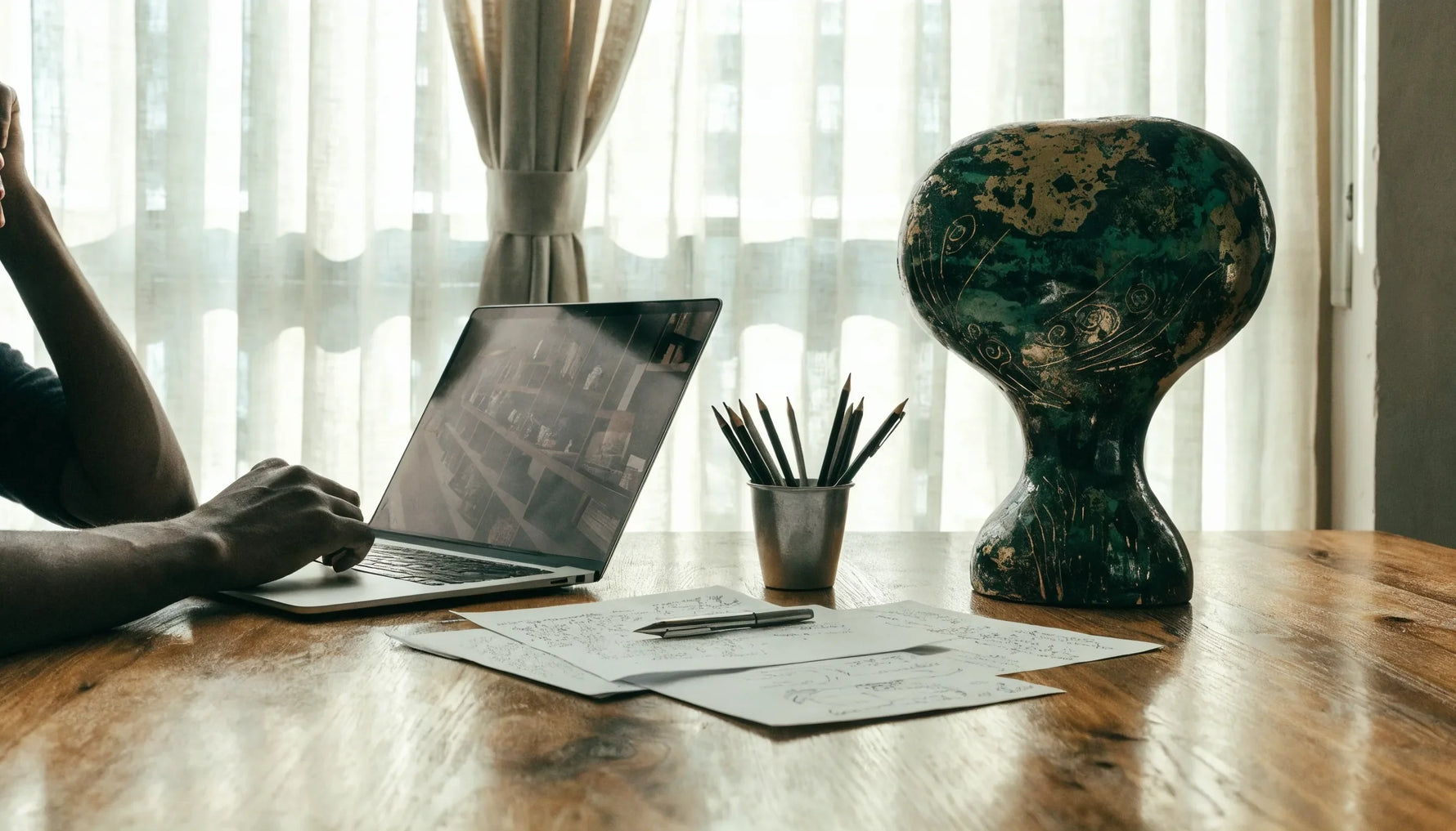 A calm research desk with an African-symbol artwork, notes and a laptop showing a soft museum archive, over-the-shoulder, no face.