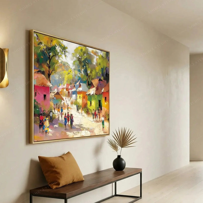 Gold-framed African village painting in a bright entryway above a wood bench with a caramel pillow and dried palm