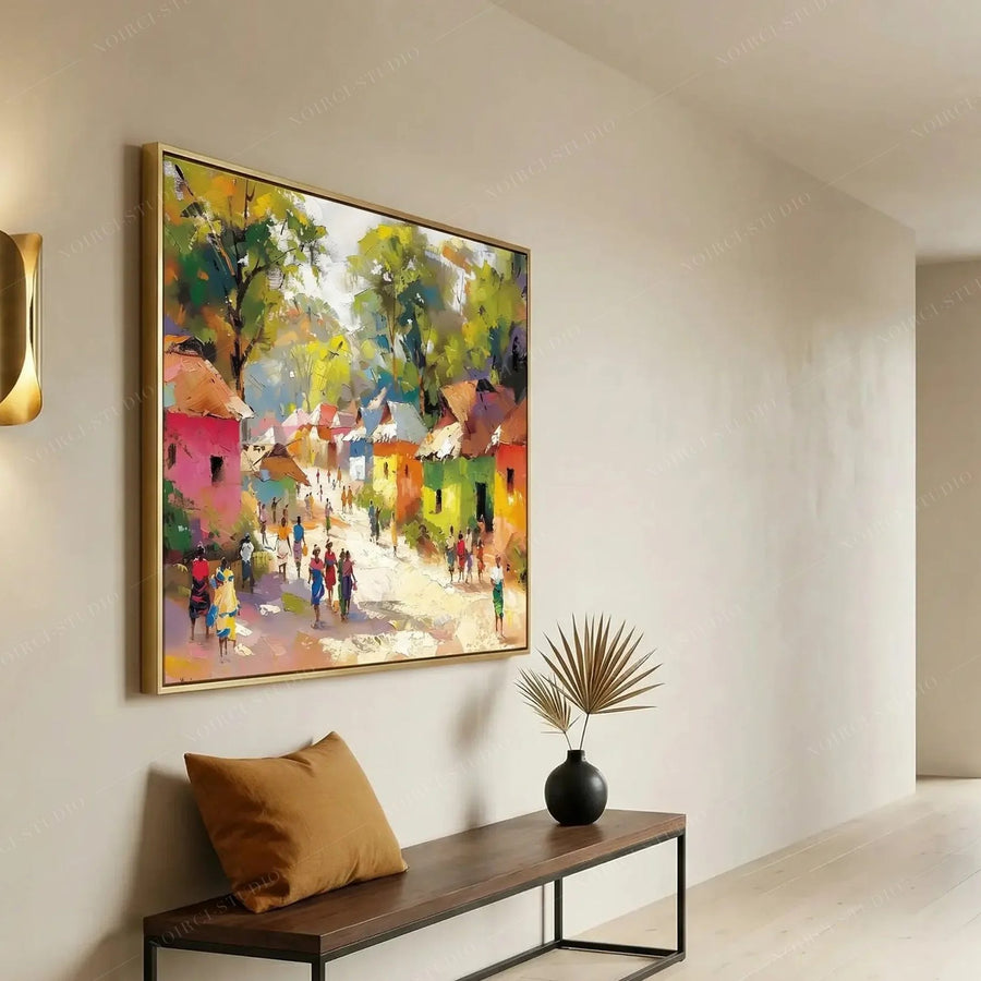 Gold-framed African village painting in a bright entryway above a wood bench with a caramel pillow and dried palm