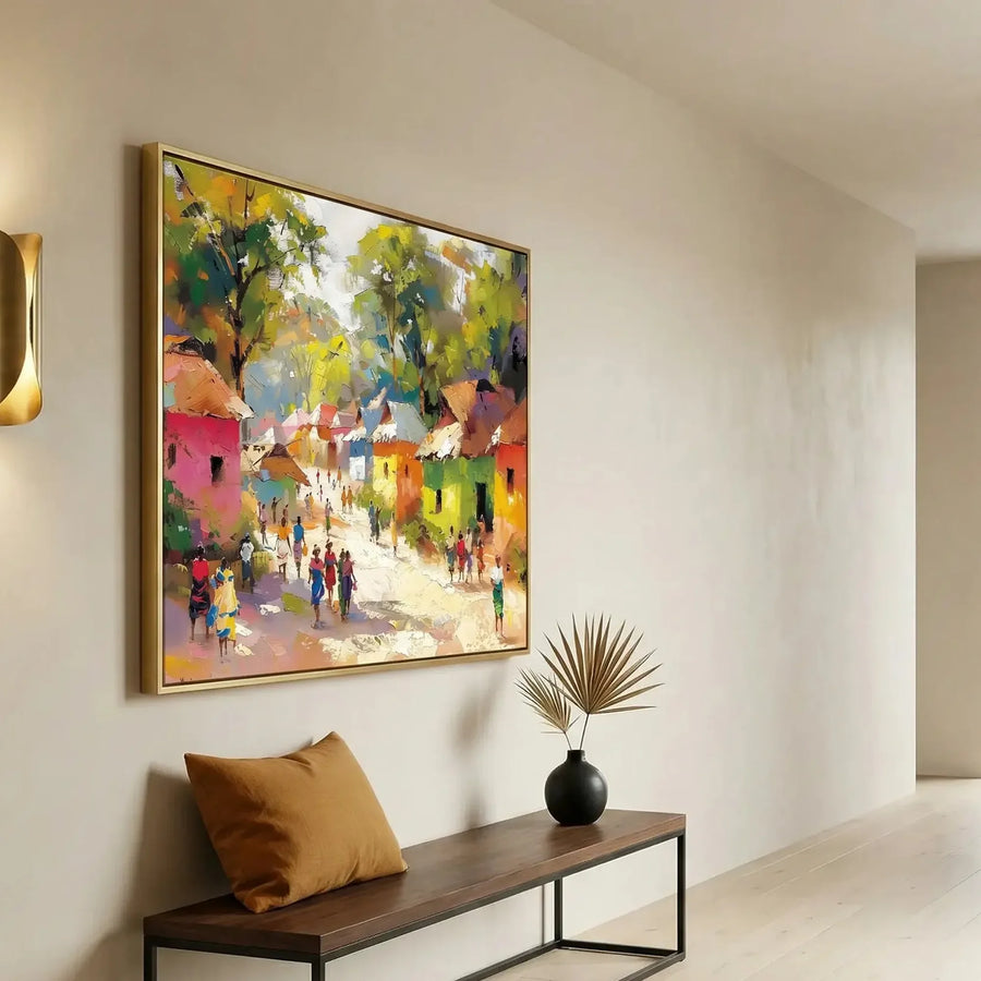 Gold-framed African village painting in a bright entryway above a wood bench with a caramel pillow and dried palm