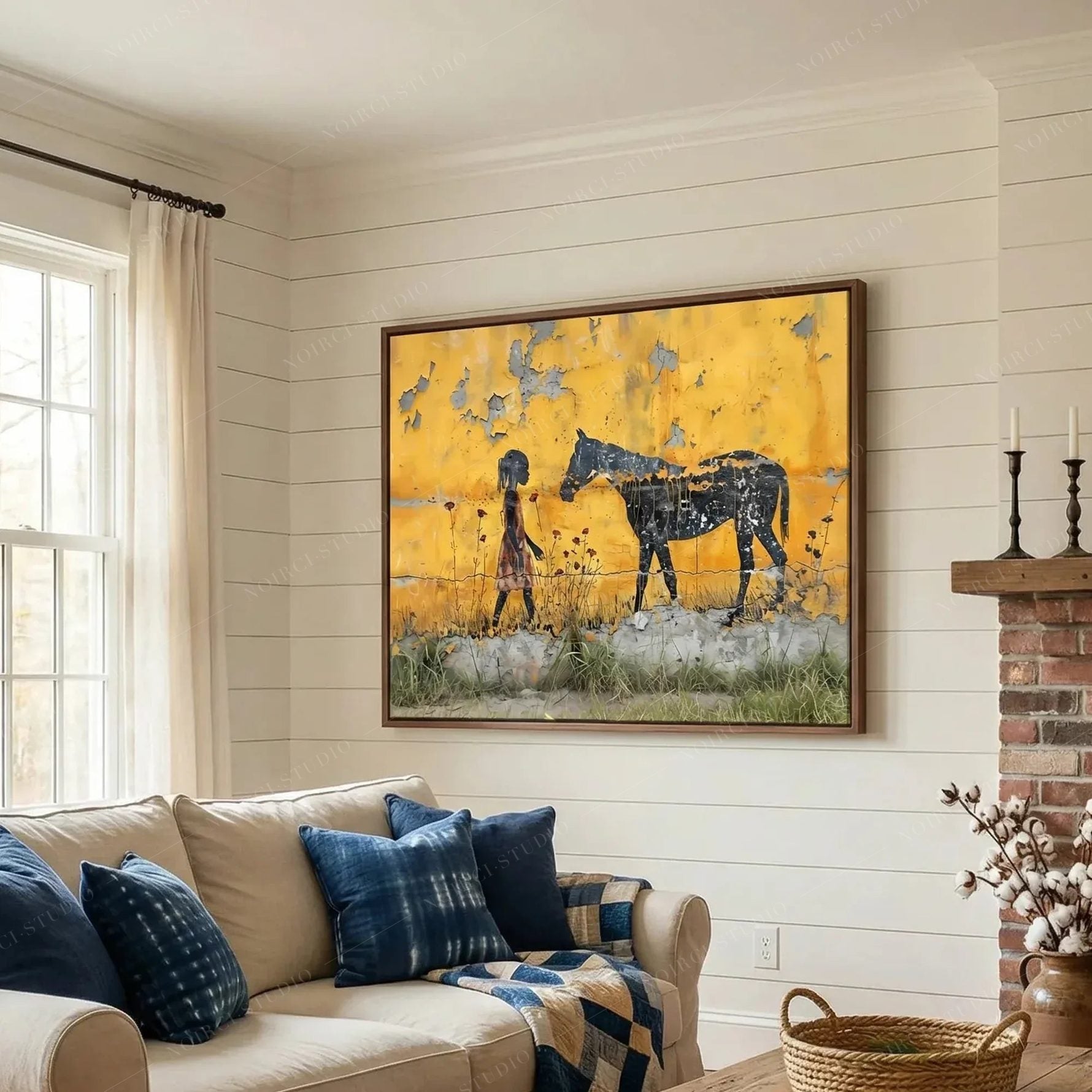 Black art for living room, framed girl and horse scene above a cream sofa with indigo pillows near a brick fireplace